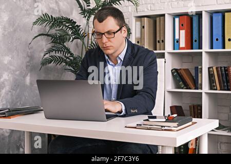 Serious, focused man in glasses and business suit is typing something using laptop while sitting in light home office on the background of rack with Stock Photo