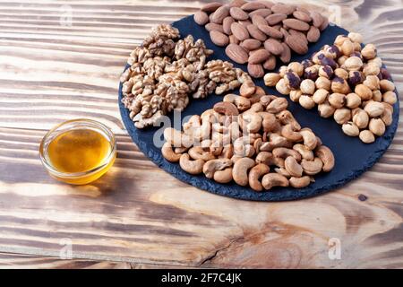 Various nuts sorted on round stone plate with honey glass bowl. Mixed nuts on wooden table. Black stone plate on wooden background. Stock Photo