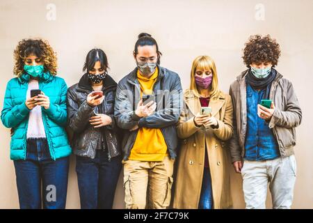 Group of young people covered by face masks using smartphone mobile device - New normal concept with friends on social media - People and technology c Stock Photo