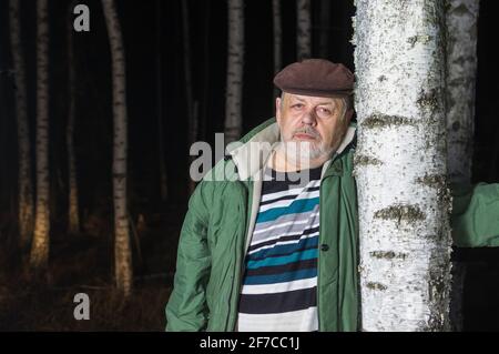 Close portrait of sad senior man leaned on birch tree in evening forest Stock Photo