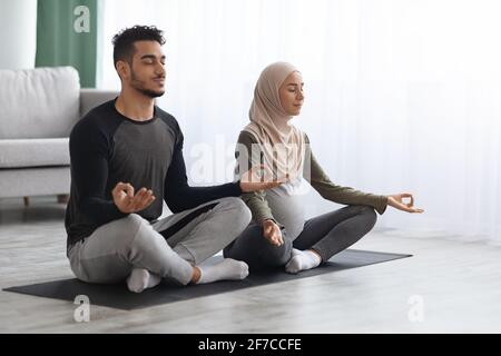 Expecting Muslim Spouses Practicing Yoga Together At Home, Sitting In Lotus Pose Stock Photo