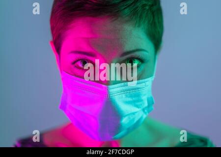 Beautiful woman with short hair wearing a face mask, looking worried and depressed in violet and green lighting. Life in quarantine. Dealing with coro Stock Photo