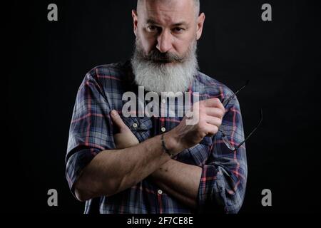 Studio portrait of mature bearded man in casual clothes with eyeglasses in hand looking directly at camera, informal lifestyle, black background. Stock Photo