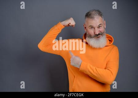 Mature bearded man dressed orange hoodie shows muscles after practising over gray studio background, look I am very strong and healthy, perfect biceps Stock Photo