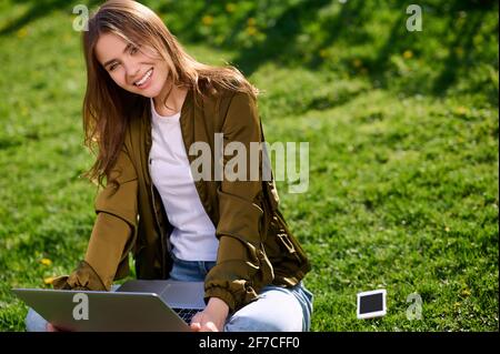 Smiling caucasian female (woman) works with laptop computer outdoors in summer park Stock Photo