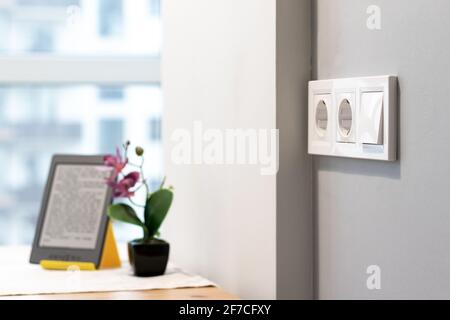 Group of white european electrical outlets and a switch located on a gray wall in a light modern kitchen with flower and e-book on the table Stock Photo