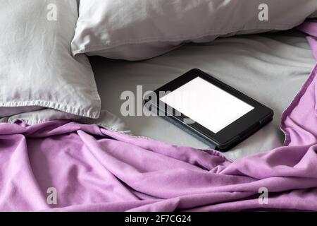 A modern black e-reader electronic book with a blank screen on a bed with two gray pillows and a lilac blanket. Mockup tablet on microfiber bedding Stock Photo