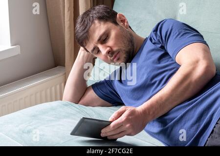 A resting man in a blue t-shirt lies on a green sofa near the window with an electronic book in one hand with the other hand under his head. Bearded m Stock Photo