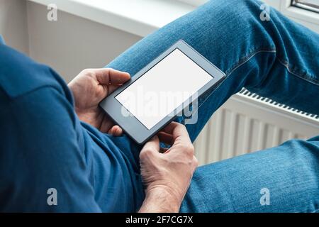 A man holds an electronic book with white blank mockup screen in his hands. Reading books at home by the window. Selective focus Stock Photo