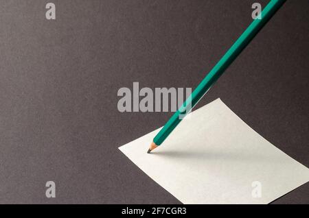 Standing pencil and square sheet of paper on a gray background. A green pencil stands on a white sticky note. Selective focus. No people Stock Photo