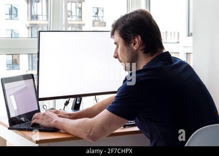A man is sitting at a workplace with laptop and a monitor near the window. Remote work from home in quarantine Stock Photo