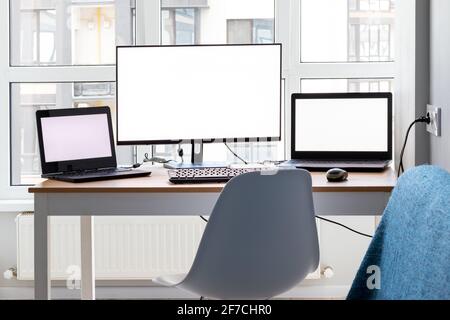 Bright workplace with two laptops and a monitor with white mockup empty screens near the window. Remote work from home in quarantine Stock Photo