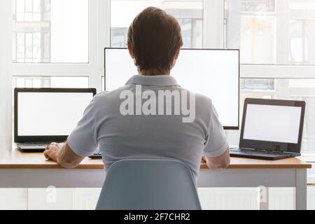 A man is sitting at a workplace with two laptops and a monitor near the window. Remote work from home in quarantine. Back view Stock Photo