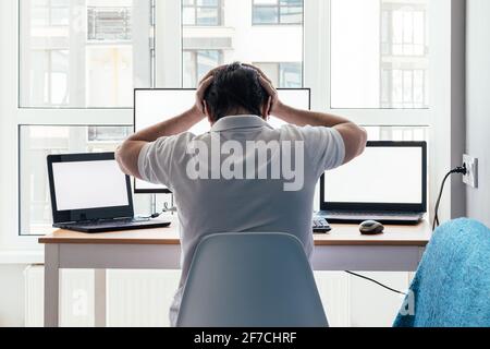 Man with hands on his head is sitting at a workplace with two laptops and a monitor near the window. Remote work from home office. Back view Stock Photo