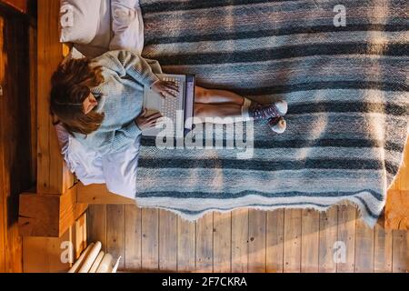 Woman in sweater and warm socks working on laptop sitting on cozy bed Stock Photo