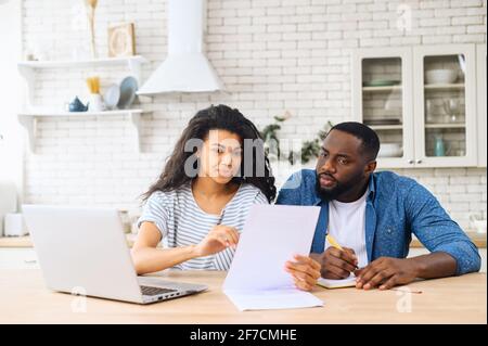 Puzzled thoughtful multiracial man and woman sitting together on table, studying documents of their family business and working with modern laptop, audit at home, finances, domestic budget concept Stock Photo
