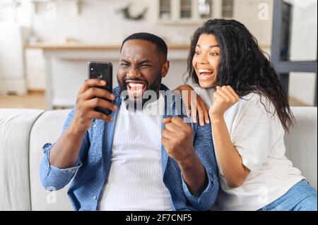 Black overjoyed couple resting on couch holding smart phone, celebrating on-line lottery win, bid betting victory moment, unbelievable opportunity or invitation, internet sale, getting prize concept Stock Photo