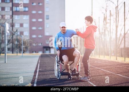 A teenage son helps his athlete father to get up from his wheelchair on an athletics track. Stock Photo