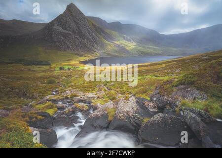 Moody, dramatic landscape of mountain stream and light rays beaming down on Tryfan peak and Llyn Ogwen and valley in Snowdonia National Park, North Wa Stock Photo