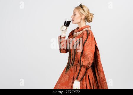 queen in dress and crown holding glass and drinking red wine on white Stock Photo