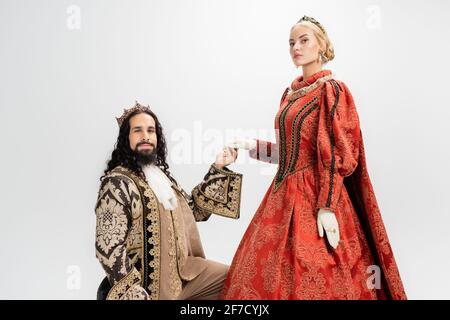 hispanic king in crown and medieval clothing holding hand of blonde queen on white Stock Photo