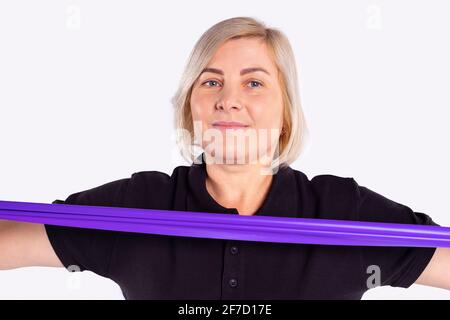 Portrait of pretty blonde senior woman exercises with violet resistance band in the gym, looking at camera. Working out and fitness, elderly activity Stock Photo