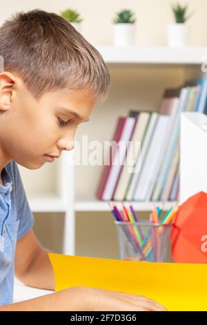 Kid create origami colored paper card white watching video tutorial on tablet computer. Education, learning, paper craft, entertainment at home Stock Photo