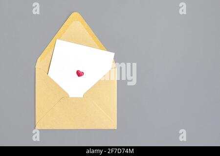 Open empty envelope made of craft paper and white leaf with small pink heart on gray background. Close-up. Top view. Copy space. Selective focus. Stock Photo