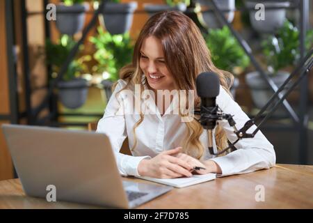Front view of smiling young beautiful blogger woman communicating with followers online by laptop. Concept of process live broadcast and talking with subscribers with modern microphone. Stock Photo