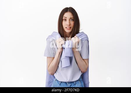 Girl looking unconfident and anxious, holding hands on sweatshirt sleeves and staring nervous at camera, have worries, scared of something, standing Stock Photo