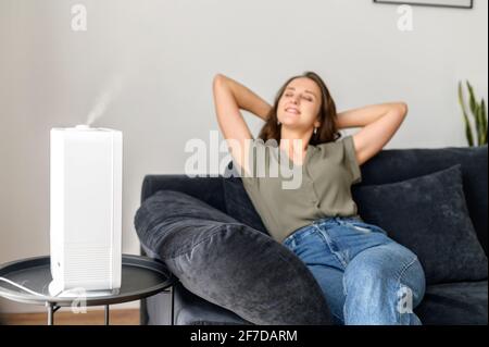 Selective focus at the humidifier, smiling and serene young woman rests on the comfortable couch under stream of fresh and cleaned air in cozy living room, housewife leaned back with eye closed Stock Photo