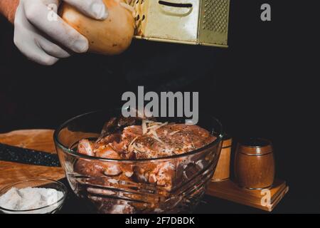 A large piece of pork is cut into pieces, marinated by the chef with spices, and the chef rubs the potatoes on a grater to the marinade.2020 Stock Photo