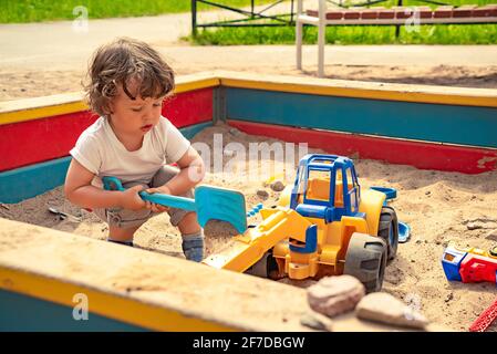 Little boy playing in the sandbox with a toy shovel and excavator outdoors on playground. Stock Photo