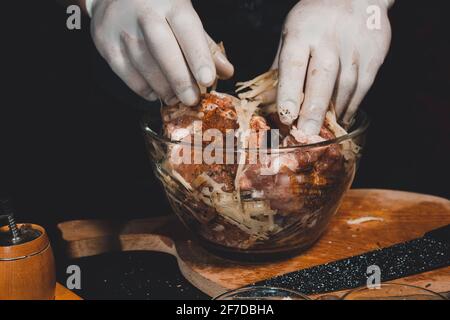 A large piece of pork is cut into pieces, marinated by the chef with spices, and the chef rubs the potatoes on a grater to the marinade.2020 Stock Photo