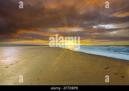 Deserted beach during sunset with orange deep blue colored clouds and low sun with sun rays on the horizon shining over the beach Stock Photo