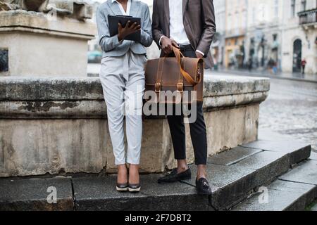 Close up of afro american woman with clipboard and young man with briefcase sitting together outdoors. Two business colleagues solving urgent working issues on street. Stock Photo