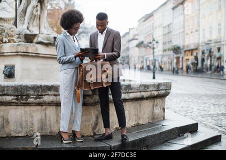 Beautiful afro american woman showing some working information on clipboard to handsome businessman in suit. Concept of cooperation and assistance. Stock Photo