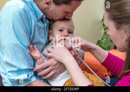 Father holding and kissing his baby boy while the mother is using the nasal aspirator to clear the mucus from the nose of her infant. Baby healthcare. Stock Photo