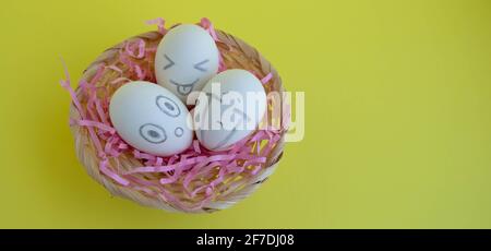On the pink tinsel in the basket are three charming white eggs with faces on a yellow background. Easter Concept. Place for your text. Stock Photo