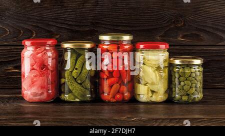 Сanned vegetables in glass jars on a dark wood shelf. Preserved artichoke in oil, pickled ginger, gherkins, capers and baby red hot peppers. Fermented Stock Photo
