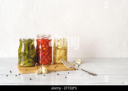 Glass jars with canned vegetables on a white wooden table. Artichoke in oil, pickled gherkins and small red hot peppers are ready to eat. Fermented. Stock Photo