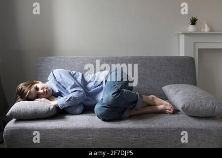 A pensive sad girl with a short hair of nineteen years in a white shirt with blue stripes looks at the camera, lies on the couch. Stock Photo