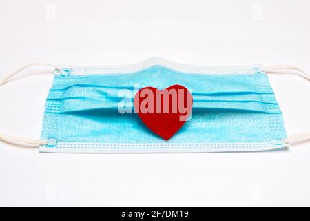 A blue medical mask with a small red felt heart on its folds, February 2021, COVID-19 Valentine's Day-themed. Stock Photo