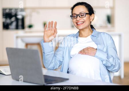 Online meeting concept. Successful pregnant mixed race woman, coworker, business lady uses laptop, communicates via video conference, conducts virtual meeting with client or employee, gesture hands Stock Photo