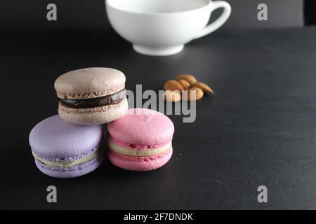 French dessert cuisine three macaroons lie pyramid to lies almonds and a cup of coffee in the background on a black background with a copyspace . Stock Photo
