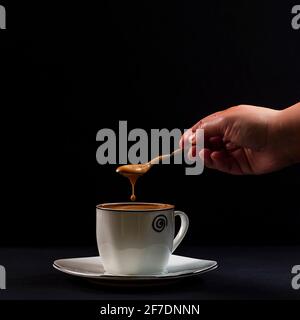 Drop of coffee with milk falling from a spoon held by a person's hand.Photo taken on a black background with artificial light in a studio.It aims to s Stock Photo