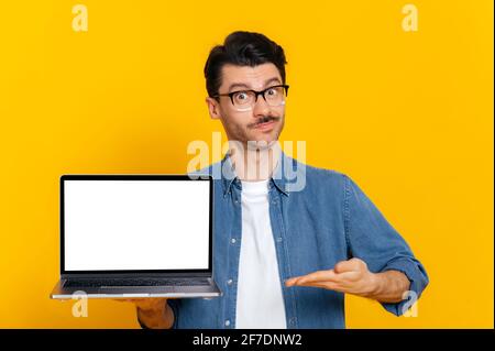 Handsome charismatic stylish caucasian guy, holding an open laptop with a blank white screen in his hand, points with his hand at it, stands on isolated orange background, place for your presentation Stock Photo