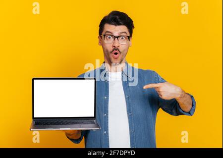 Amazed surprised cheerful caucasian unshaven guy with glasses and denim shirt holds open laptop with blank white screen, points finger at it, standing on isolated orange background Stock Photo