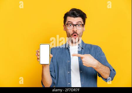 Amazed shocked surprised caucasian guy in denim shirt and glasses holds smartphone with blank white screen for your text and points finger at it, stands on isolated orange background, looks at camera Stock Photo