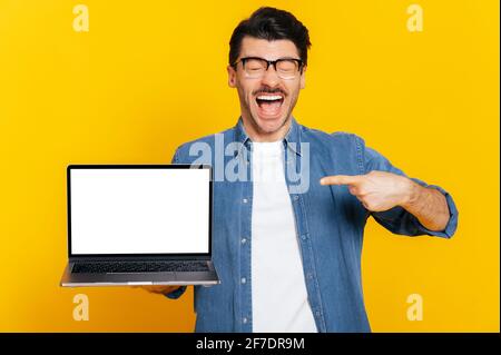 Happy shouting caucasian excited stylish guy with glasses, holds an open laptop with empty white screen, points finger at it, standing on isolated orange background, smiling friendly Stock Photo